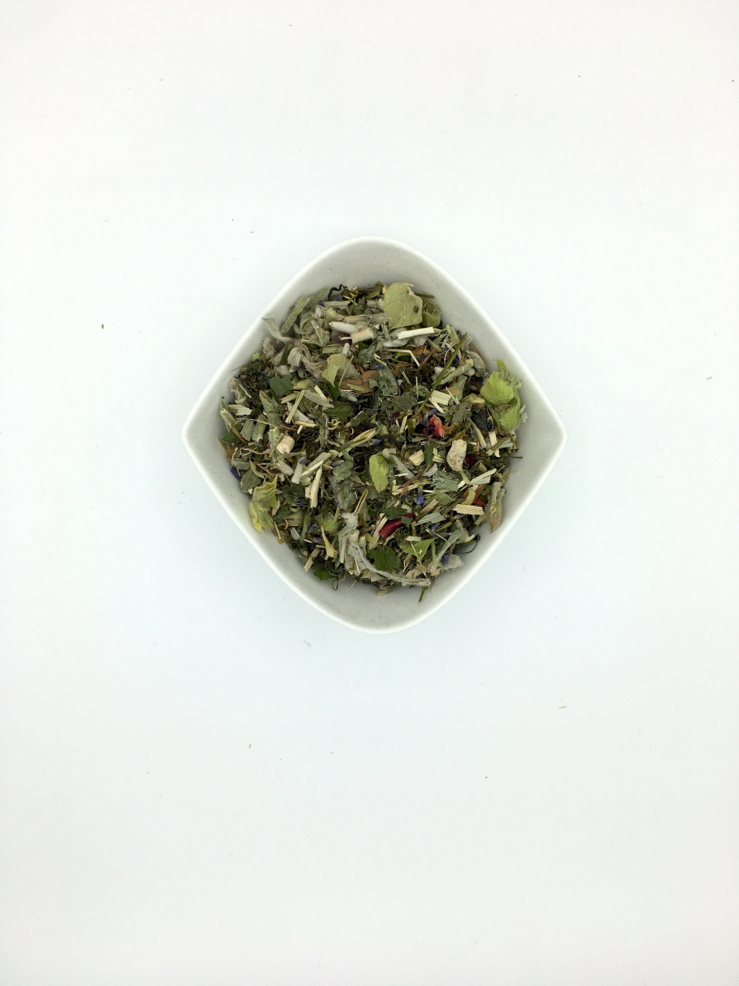 Die weiße, rautenförmige Porzellanschale enthält dunkel- bis hellgrüne, mittelgrob geschnittene Kräuter mit größeren hellgrünen Blütenstücken Griechischen Bergtees und ein paar Rosenblütenblättern.