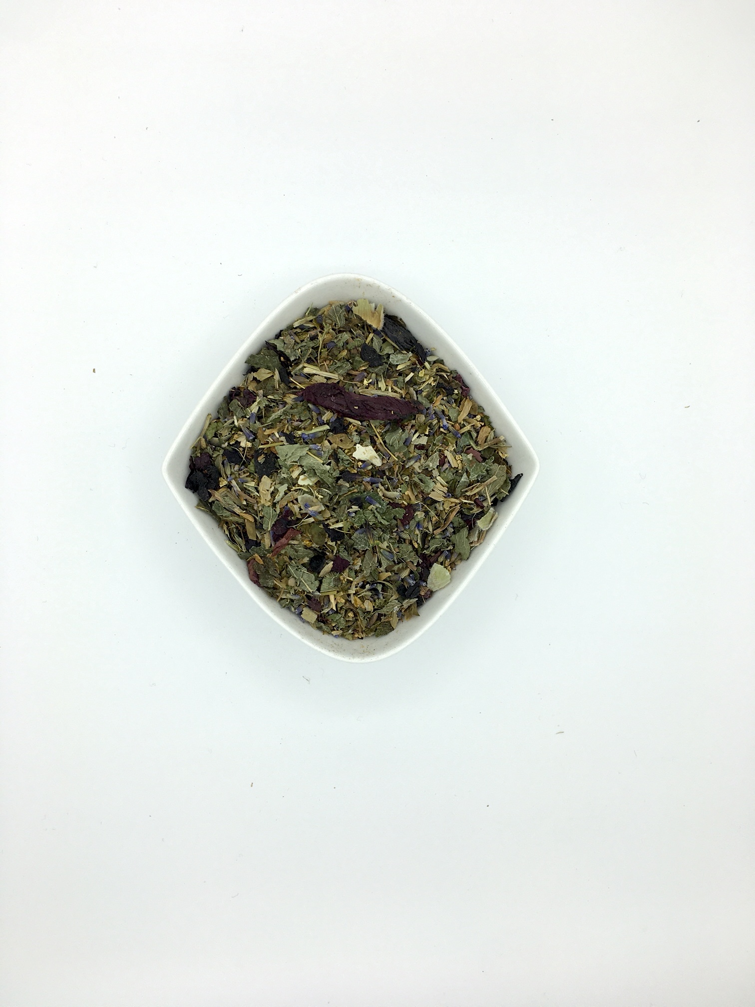 Die rautenförmige, weiße Porzellanschale enthält mittelgrob geschnittene, dunkelgrüne Kräuter mit braungelben Hopfenblütenblättern, tiefvioletten Stockrosenblüten und dunkelroten Klatschmohnblüten.