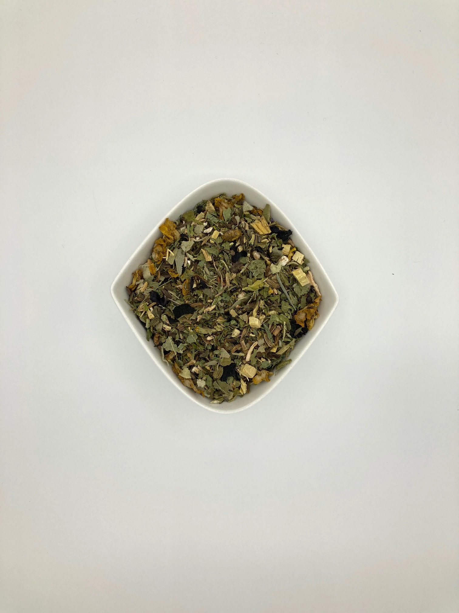 Die weiße rautenförmige Porzellanschale enthält dunkelgrüne Blattstückchen und gelbe Blüten und helle Wurzelstücke.