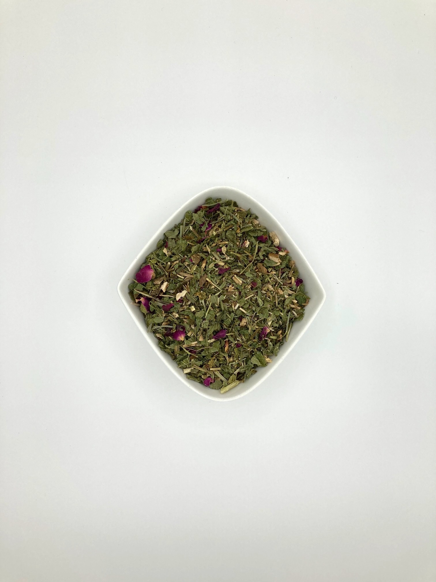 Die weiße rautenförmige Porzellanschale enthält vorwiegend mittelgrüne Blattfragmente, gelblbräunliche Wurzelstücke und einige Rosenblütenblätter.